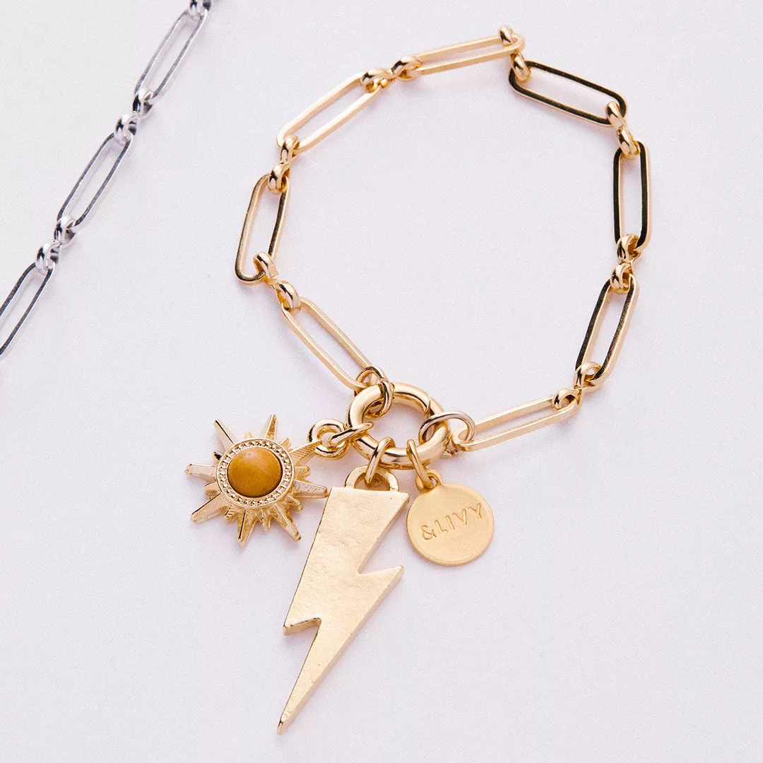 &LIVY Paperclip Intention Chain Bracelet in finely finished gold close-up of paperclip links with charm holder featuring sun charm with amber gemstone lightning bolt charm and branded coin charm with silver bracelet visible in background