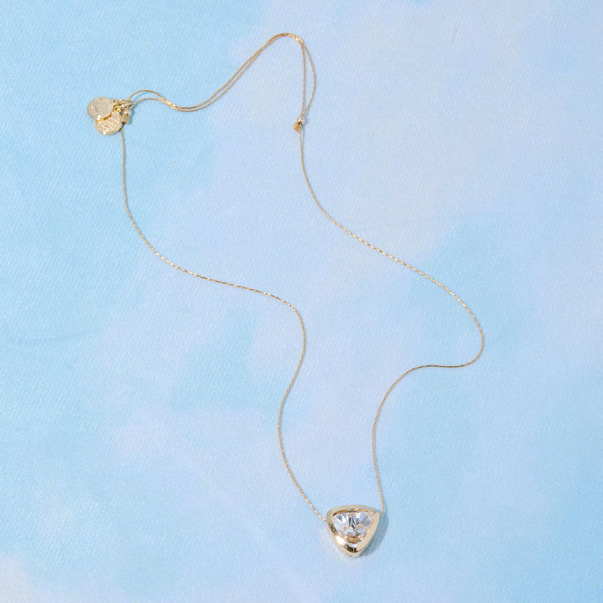 &LIVY Let The Light In Pendant Necklace in finely finished gold full length view showing clear crystal heart pendant with gold bezel setting and small heart charm detail on delicate gold expandable chain against light blue background