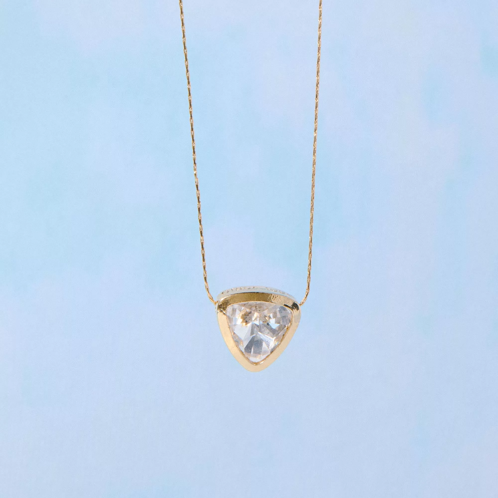 &LIVY Let The Light In Pendant Necklace in finely finished gold featuring large clear crystal pendant in heart shape with gold bezel setting on delicate gold chain against blue sky background