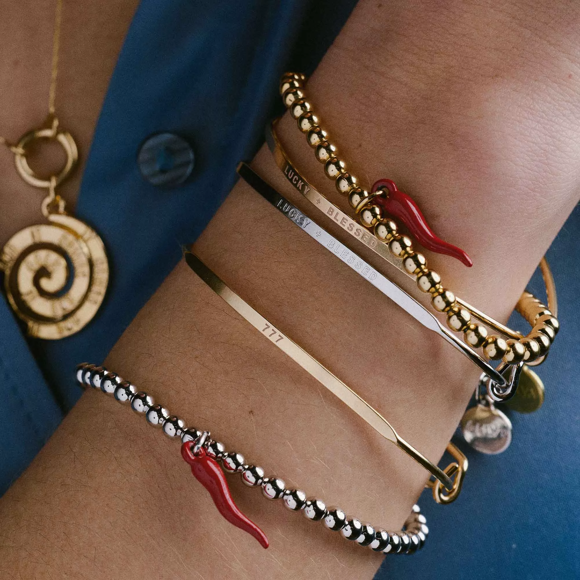 &LIVY bracelet stack worn on wrist featuring Lucky + Blessed Posy bangle with gold beaded bracelets, red Italian horn charms, and 777 engraved bangle