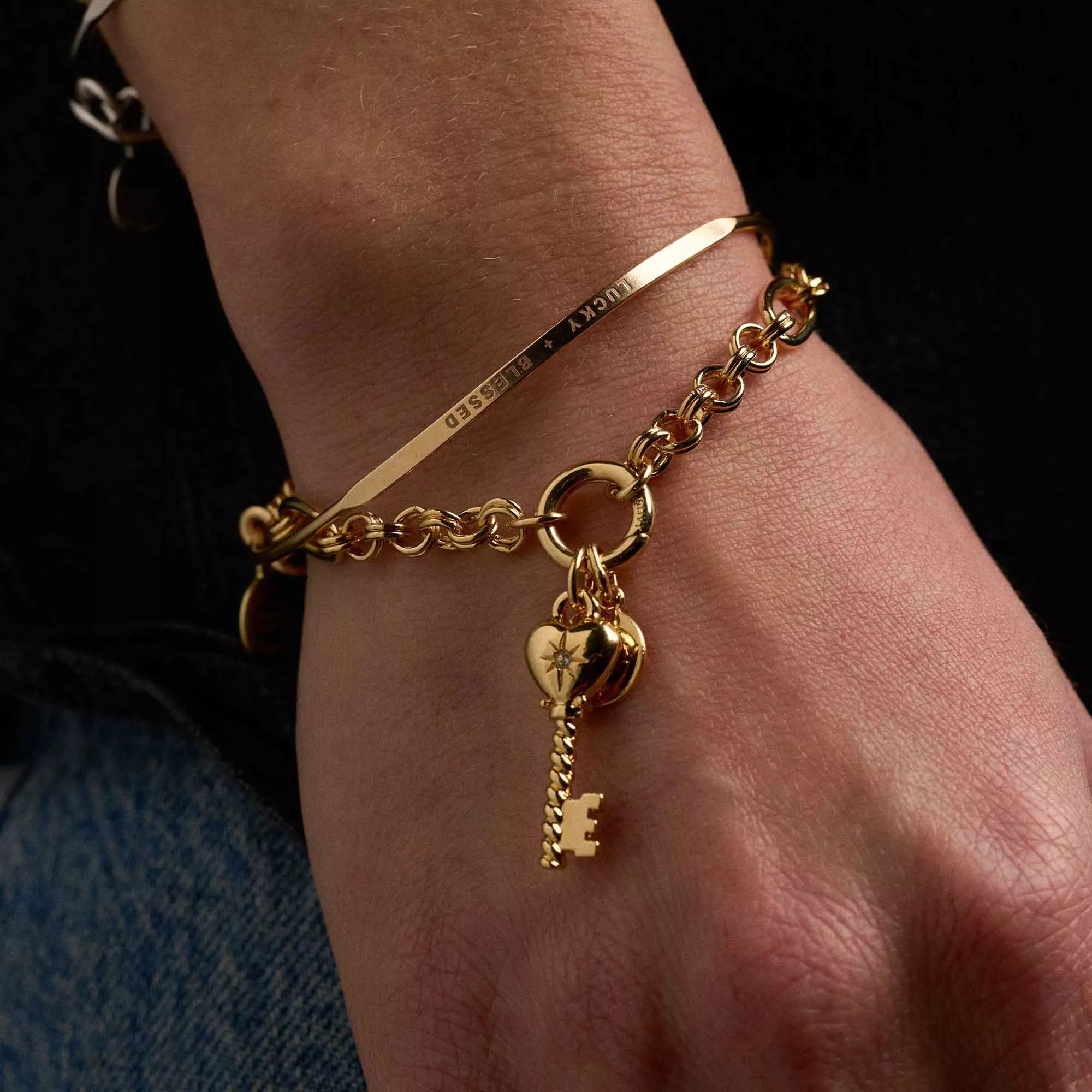 &LIVY Key To My Heart Charm Bracelet in finely finished gold worn on model wrist showing engraved Lucky and Blessed bar charm heart lock with gemstone key charm and link chain